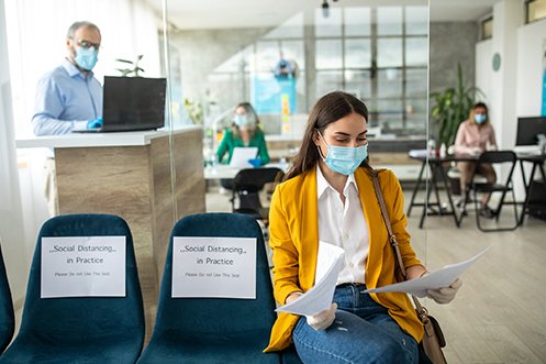 Four employees working, one man standing at a counter, one woman sitting in a chair, two women sitting at separate desks, all are wearing masks in a socially distant setting reflecting new workspace configurations to limit COVID-19 issues