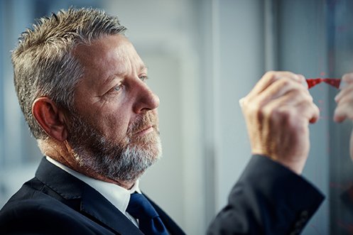 Business owner with mustache and beard, wearing dark blue suit and dark blue tie, facing right, holding a red pen while he is marking up operational charts as part of post-pandemic business planning