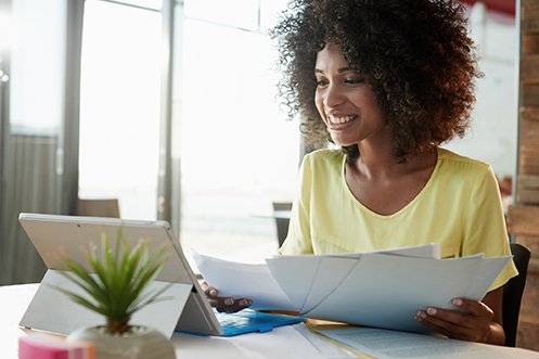 Woman smiling, with dark hair, holding paperwork in front of a computer, reviewing her company’s 2020-21 Corporate Relocation Budget