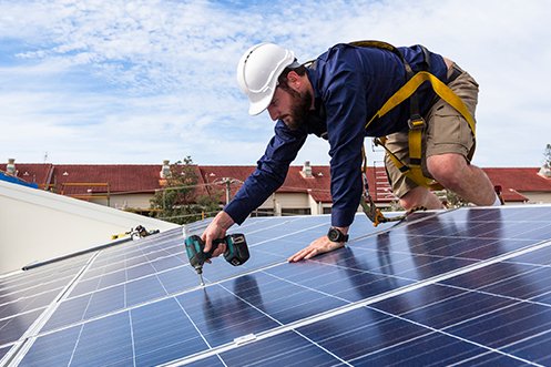 Solar technician wearing blue shirt, tan shorts, white hard had, with dark hair and beard, on roof of home installing solar panels to meet the requirements of the California solar panel mandate