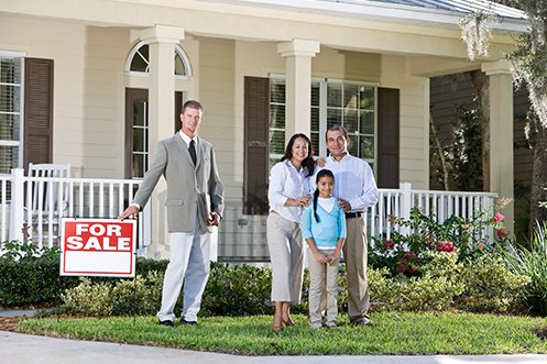 Licensed appraisal professional wearing a tan jacket and white pants, ready to prepare a Mortgage Appraisal in front of a home that is for sale, three family members husband, wife, and daughter who want to buy the home are standing in front of home smiling