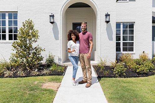Woman in white shirt with dark curly hair, many in red shirt and tan pants, blonde/brown hair and full beard, young couple in front of house holding keys, happy they looked at renting versus buying and chose to buy this beautiful home