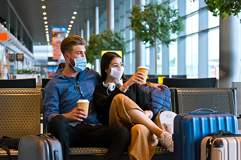 Man and woman sitting in airport, both wearing masks, both holding coffee cups, man is wearing a blue shirt and black pants, woman is wearing a black shirt and tan pants, both are looking out the window, following COVID-19 travel recommendations