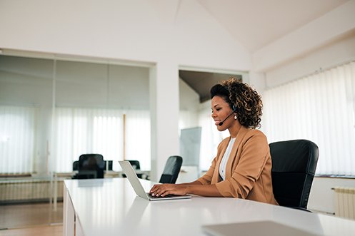 Woman with dark hair and wearing a tan jacket, sitting at a table in front of a computer with headset, conducting a personalized webinar for an initial consultation and needs assessment as part of virtual destination services