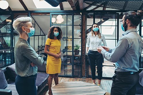 : Four coworkers wearing masks in a shared office space, three women and one man, talking about the top 5 COVID-19 risks for their company’s relocation policy