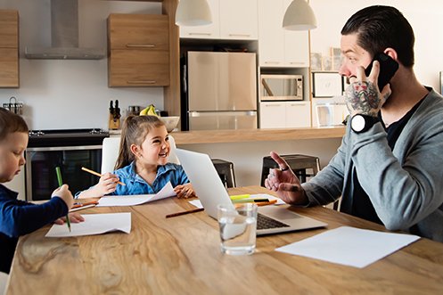 Man with dark hair and beard, wearing a gray sweater and large wristwatch, talking on a cell phone, sitting at kitchen counter, working on a laptop computer, working from home with kids, one boy and one girl sitting across from him, smiling