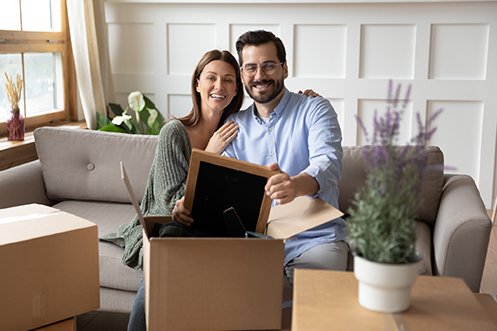 Woman with long dark hair and wearing a green sweater, man with dark hair, mustache, and beard wearing glasses and a blue shirt, both sitting on a couch, smiling, unpacking a box in their new home after their relocation mortgage application was approved