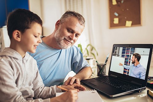Father with mustache and beard, wearing a blue shirt, son with dark hair, wearing a gray pullover, both watching a virtual tutoring session on a computer, as the father helps the son with the lesson