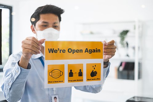 Man with dark hair, wearing a face mask, hanging a sign in a window saying the business is open again as employees return to workplaces safely