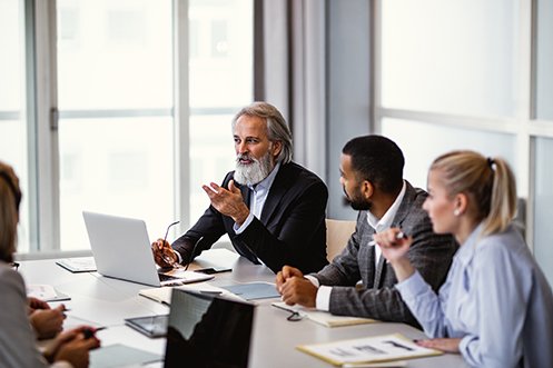 Man with gray hair and beard, wearing jacket, in front of open laptop, holding eyeglasses and gesturing with one hand; man next to him, with dark hair, wearing a jacket, looking at the man with gray hair; woman with blonde hair wearing blue shirt, holding a pen; all are Relocation Department team members sitting at a meeting table in a conference room, all wearing business professional attire, planning future global relocations and discussing pre-decision tax planning to reduce global tax obligations