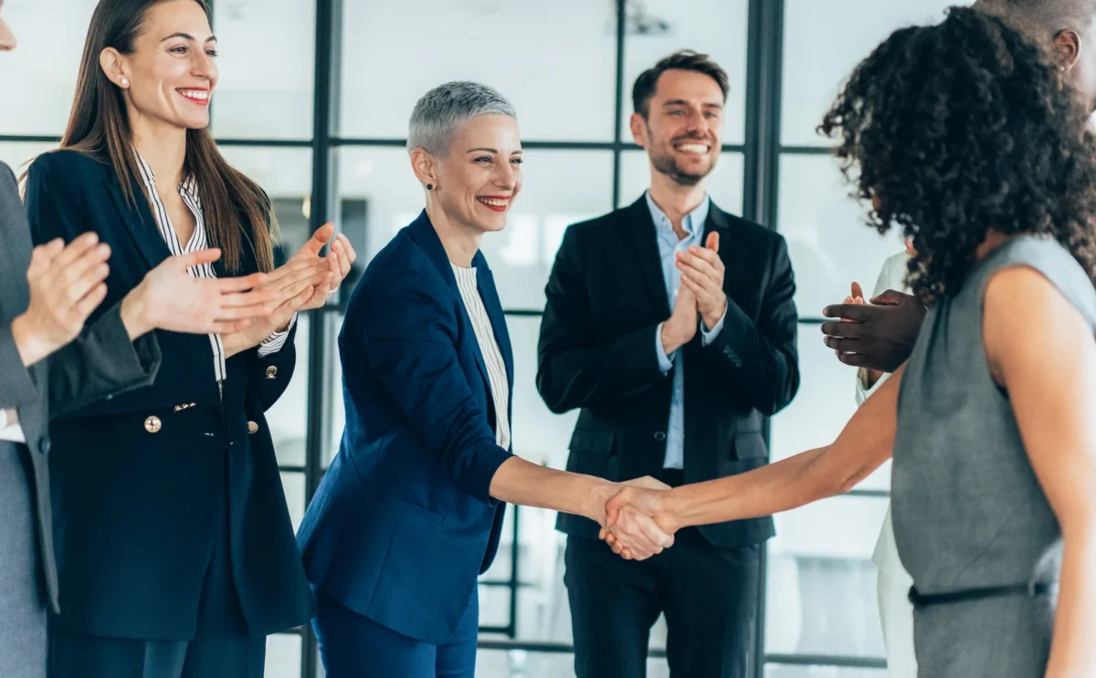 Group of business people at the office, two of them shaking hands for successful deal, colleagues around applauding