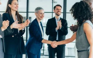 Group of business people at the office, two of them shaking hands for successful deal, colleagues around applauding