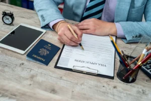 Close-up of male hands filling out visa documents at a table. On the table there is an American passport and an American flag